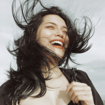 A woman with long black hair beams with joy outdoors under a cloudy sky, wearing a black top and holding the strap of her Cultivating Joy bag as the wind playfully tousles her hair.